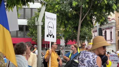 Grupo-De-Personas-De-Diferentes-Orígenes-Reunidos-Pacíficamente-En-La-Plaza-De-Brisbane-Expresan-Públicamente-Su-Ira-Sosteniendo-Un-Cartel-Que-Muestra-A-Vladimir-Putin-Como-El-Terrorista-Número-Uno