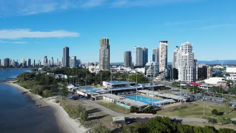 Creative-drone-view-of-a-inner-city-Aquatic-Centre-on-a-coastal-strip-with-a-towering-skyline-backdrop
