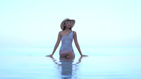 Classy-Young-Woman-in-Swimsuit,-Hat-and-Sunglasses-Sitting-on-Infinity-Pool-Edge-With-Endless-Tropical-Sea-Horizon-in-Background,-Full-Frame