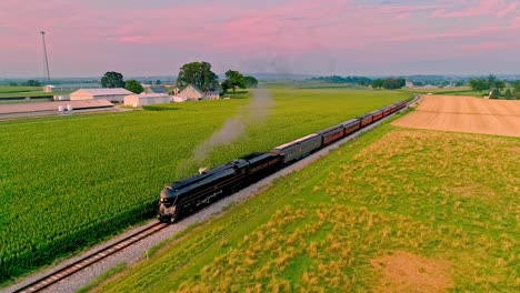 Una-Vista-Aérea-De-Un-Tren-De-Pasajeros-De-Vapor-Antiguo-Que-Sopla-Humo-Y-Vapor-Que-Viaja-A-Través-De-Campos-De-Maíz-Fértiles-Durante-La-Hora-Dorada-En-Un-Día-Soleado-De-Verano