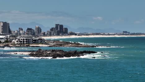 Rocky-shore-and-buildings-in-Small-Bay-beach,-Cape-Town,-South-Africa,-with-Bloubergstrand-in-background