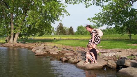 Eine-Vergrößerte-Aufnahme-Von-Zwei-Kindern,-Die-Auf-Einem-Felsen-Am-Ufer-Eines-Sees-Sitzen,-Und-Ihre-Mutter-überwacht,-Wie-Sie-Hinter-Ihnen-Steht