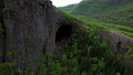 Aerial-flyover-of-a-hidden-cave-in-the-mountain-with-people-standing-next-to-the-entrance-in-Helgeland,-Norway