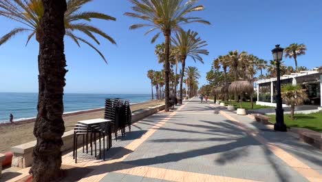 Caminando-Por-Un-Sendero-Tropical-Junto-Al-Mar-En-Un-Día-Soleado-Con-Cielo-Azul-Y-Palmeras-En-Marbella-España,-Destino-De-Vacaciones-Paseo-Marítimo-San-Pedro-De-Alcantara-Naturaleza,-Tiro-4k