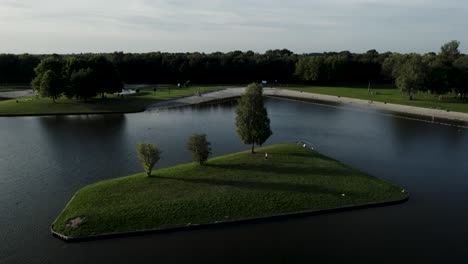 Aerial-rotate-pan-around-single-grass-island-with-three-trees-and-sunset-shadow-in-recreational-pond-in-The-Netherlands-seen-from-the-air-with-landscaped-environment-including-man-made-beach