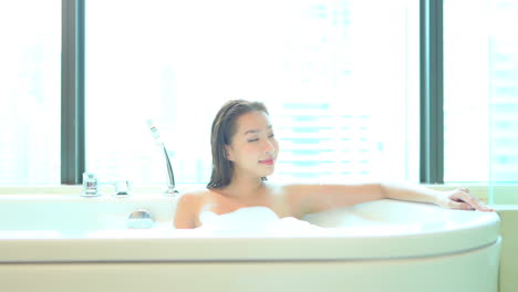 An-Asian-woman-comfortably-taking-bath-is-lying-in-a-bathtub-full-of-foam-at-luxury-apartment-complex-with-Glass-walls