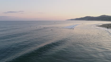 Tracking-aerial-following-wave-in-as-surfers-ride-to-shore-at-sunrise,-4K