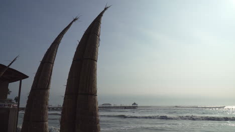 Athentic-Caballito-de-totora-Boats-Along-the-Huanchaco-Beach-in-Trujillo,-La-Libertad,-Peru-with-Muelle-de-Huanchaco-Pier-in-the-Background