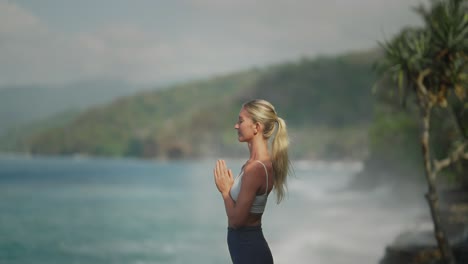 Modelo-Femenino-De-Yoga-Rubio-En-Forma-De-Pie-En-Una-Relajante-Meditación-Zen-Con-Poderosas-Olas-Rompiendo-En-La-Costa-De-Bali,-Slowmo-120fps