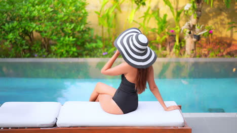 Back-to-the-camera,-a-young-woman-wearing-a-one-piece-bathing-suit-and-a-huge-matching-straw-sun-hat-turns-to-look-at-the-swimming-pool