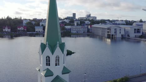 Flying-past-bell-tower-of-Free-Church-in-Reykjavik,-Tjörnin-lake,-aerial