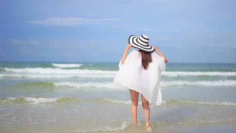 Lonely-Woman-on-Sandy-Beach-Running-in-Fluttering-Tunic-and-Floppy-Hat,-Raising-Hand-Full-of-Joy-in-Front-of-Tropical-Sea-Waves,-Back-View,-Selective-Focus,-Full-Frame