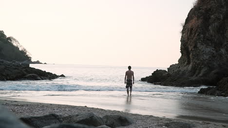 Man-slowly-walks-into-ocean-in-small-peaceful-cove-at-sunset,-slow-motion
