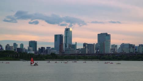 Han-river-Waterfront-with-Skyscrapper-Buildings-and-Sporty-Tourists-Paddling-on-Paddleboards,-Kayaking-and-Windsurfing-During-Sunset-In-Seoul,-Jamsil-District,-South-Korea