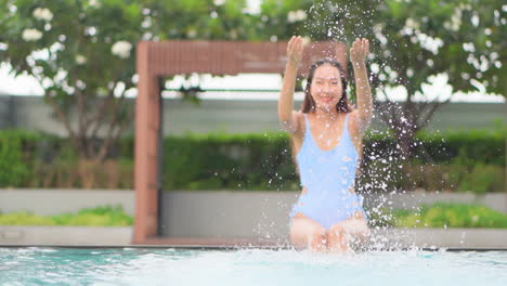 Happy-Asian-woman-in-blue-swimwear-splash-water-with-hands-while-sitting-on-the-border-of-the-swimming-pool,-slow-motion
