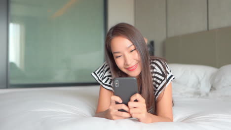 Happy-Asian-woman-typing-a-message-on-mobile-phone-using-both-hands-while-comfortably-lying-on-a-big-bed-in-hotel-room