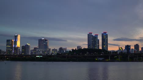Seoul-Night-Cityscape-From-Waterfront-of-Han-River,-Cars-Traffic-on-Olympic-daero-Highway,-Trade-Tower,-and-Asem-Tower-on-Purple-Sky-at-Sunset,-Yacht-Pass-by,-People-do-Water-Sports