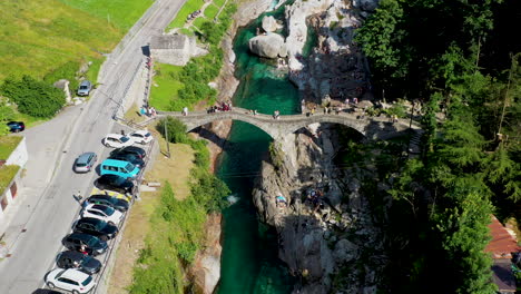 Drone-shot-of-man-jumping-off-Ponte-dei-Salti-the-double-arched-medieval-stone-pedestrian-bridge-over-the-clear-water-of-the-Verzasca-River