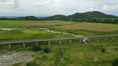 Drone-footage-sliding-towards-the-right-angled-revealing-the-elevated-railway,-farmland-with-cattle,-mountains-and-lake-in-the-horizon,-Muak-Klek,-Saraburi,-Thailand