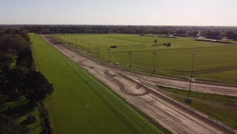 Aerial-flyover-empty-horse-racetrack-during-sunny-day-in-Buenos-Aires---Green-grass-field-surrounded-by-sandy-track