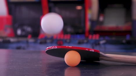 Roter-Tischtennisschläger,-Der-Auf-Einem-Orangefarbenen-Ping-Pong-Ball-Liegt,-Mit-Einem-Weißen-Springenden-Ball-In-Einem-Vergnügungsspiel-Arcade-Raum