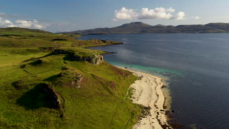 Rotating-drone-shot-of-Coral-Beach-in-Claigan-with-white-sandy-beaches,-tropical-blue-water-and-mountains-in-the-distance,-in-Scotland
