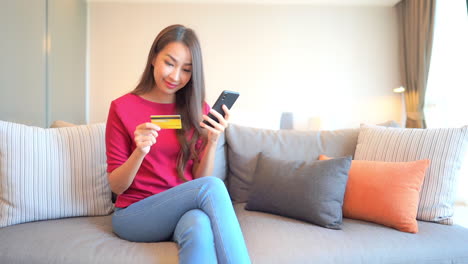 Junge-Asiatische-Frau,-Die-Auf-Sofa-Sitzt-Und-Kreditkarte-Und-Smartphone-Hält