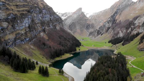 Aerial-flyover-away-from-Seealpsee-in-Appenzell,-Switzerland-with-pan-down-motion-from-the-Alpstein-peaks-towards-the-reflection-on-the-lake