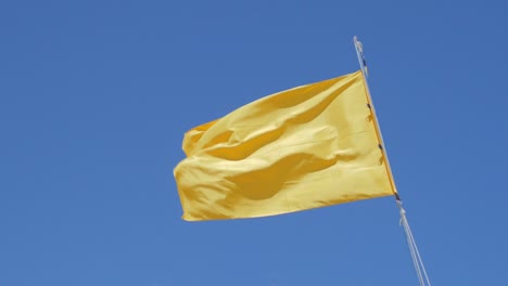 Yellow-flag-waving-in-the-wind-against-clear-blue-sky,-slow-motion-low-angle-shot