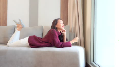 A-pretty-young-woman-in-an-oversized-sweater-and-slacks-lies-on-a-couch-on-her-stomach,-daydreams-while-looking-out-the-window