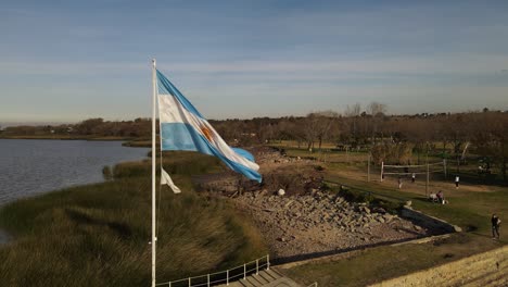 Slow-motion-track-shot-of-waving-Argentinian-flag-waving-in-wind-during-sunset---beautiful-park-and-sea-shore-in-background