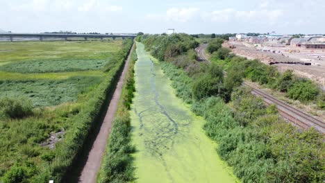 Green-algae-covered-countryside-canal-alongside-new-housing-build-site-aerial-view-rising-shot