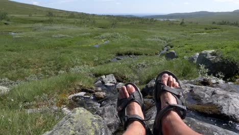 An-adult-male-resting-his-feet-next-to-spring-water-flowing-through-rocks-and-enjoy-beautiful-nature-on-green-grass-mountain-during-summer-at-Jamtland-Sweden