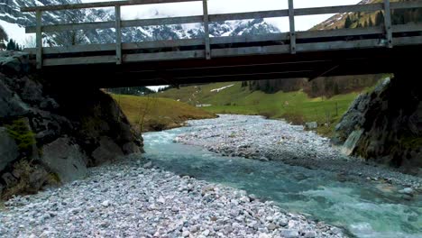 Close-low-aerial-drone-fly-through-flight-below-a-bridge-at-scenic-Ahornboden-Engtal-valley-along-Rissach-mountain-river-in-the-Bavarian-Austrian-alps-on-a-cloudy-and-sunny-day-in-nature