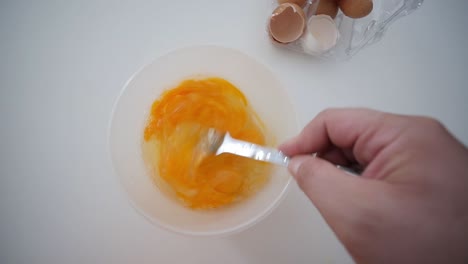 Whisking-Egg-Yolks-With-Fork-In-Bowl-Top-View-With-Broken-Egg-Shells-In-Background