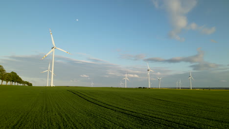 Sunrise-at-wind-turbine-park-with-green-field-and-moon-in-sky,-aerial