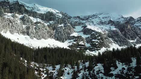 Wide-panorama-flightup-a-glacier-rock-snow-mountain-top-near-Bavaria-Elmau-castle-in-the-Bavarian-Austrian-alps-on-a-cloudy-and-sunny-day-along-trees-and-forest-in-nature-with-avalanches-going-down