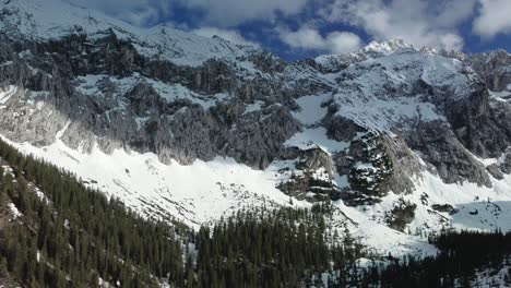 Panorama-descend-flight-at-a-glacier-rock-snow-mountain-top-near-Bavaria-Elmau-castle-in-the-Bavarian-Austrian-alps-on-a-sunny-day-along-trees-and-forest-in-nature-with-avalanches-going-down