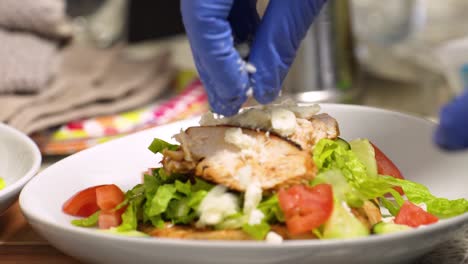 Closeup-of-cook's-gloved-hands-crumbling-feta-cheese-over-a-grilled-chicken-salad-in-a-shallow-bowl