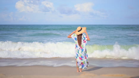 Woman-in-colorful-wrap-and-large-hat-walks-toward-ocean-and-lets-water-wash-over-her-legs-while-looking-out-to-sea