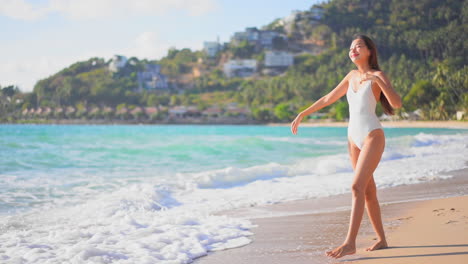 Happy-asian-woman-in-swimsuit-walking-on-sandy-beach-by-sea-waves,-raising-hands-full-of-joy,-slow-motion
