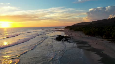 Aerial-of-sunset-reflecting-in-the-water-with-waves-and-surfers-in-the-pacific-ocean-in-Tamarindo,-Costa-Rica