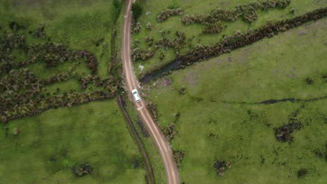 Top-down-aerial-view-of-white-car-driving-down-winding-gravel-road-and-over-stream-surrounded-by-vibrant-green-grassy-plains