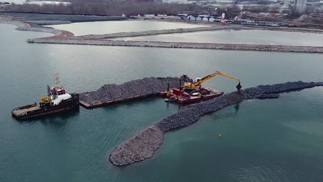 Excavator-on-barge-building-breakwater-from-rocks-near-waterfront-port