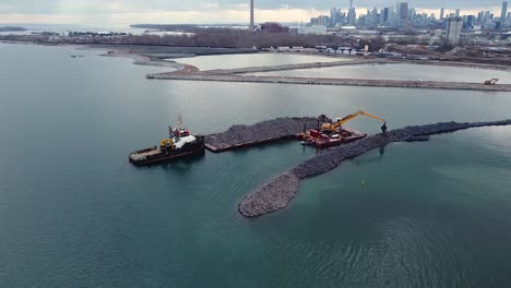 Excavator-on-barge-building-breakwater-from-rocks-with-Toronto-skyline-in-background