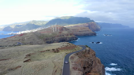 Smooth-drone-footage-of-a-car-driving-along-a-coastal-road-with-huge-cliffs-and-views-out-to-sea,-with-a-view-of-the-island-of-Madeira,-Portugal