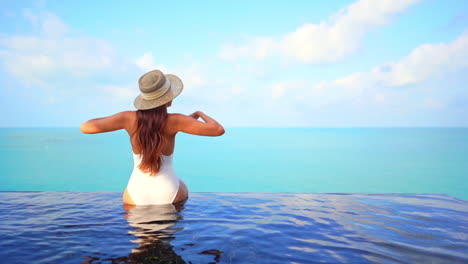 Female-on-tropical-vacation,-sitting-on-infinity-pool-border-and-raising-hands,-turquoise-caribbean-sea-and-stunning-horizon-in-background