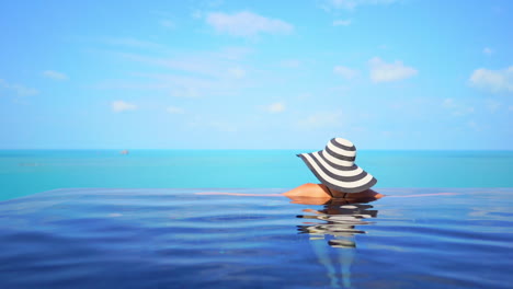 Back-of-woman-in-infinity-swimming-pool-with-summer-hat-enjoying-on-horizon-view-above-turquoise-caribbean-sea-on-sunny-summer-day,-slow-motion