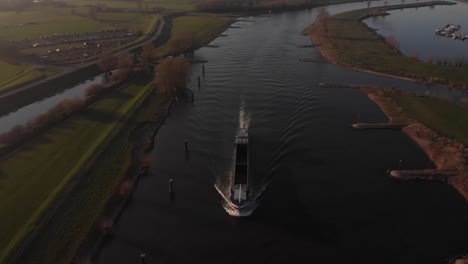 Backwards-aerial-reveal-of-picturesque-floodplain-landscape-along-the-river-IJssel-with-a-large-cargo-ship-leaving-a-wrinkled-trail-in-the-water