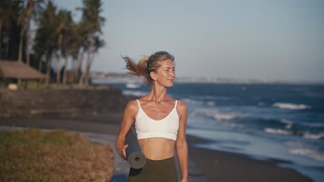 Atractiva-Mujer-Sosteniendo-Colchoneta-De-Yoga-Caminando-Por-La-Orilla-Respirando-Aire-Fresco,-Bali